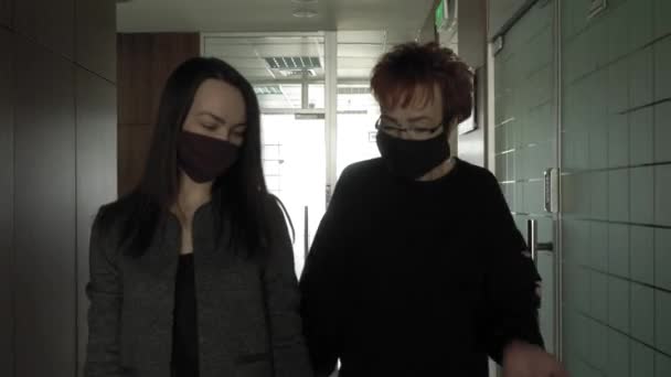 Trabajo de oficina durante una pandemia COVID-19. Joven mujer de negocios y una anciana con máscaras protectoras hablando a lo largo del pasillo de la oficina . — Vídeo de stock