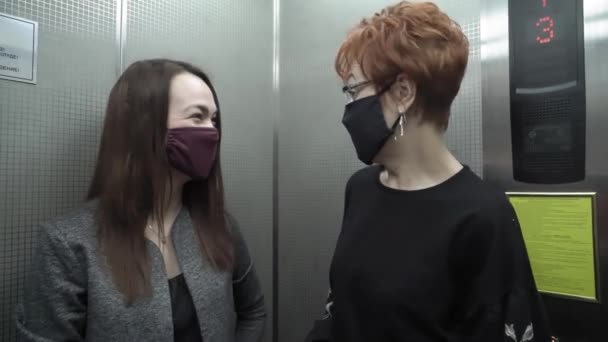 Irodai munka egy világjárvány idején COVID-19. Egy fiatal üzletasszony és egy védőruhás idős nő beszélgetnek a liftben.. — Stock videók