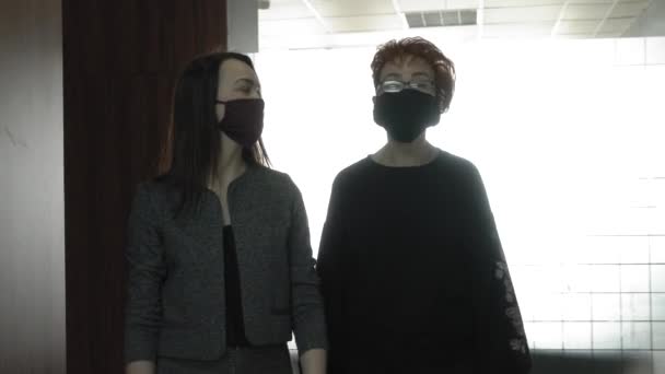 Salgın bir COVID-19 sırasında ofis işi. Genç bir iş kadını ve koruyucu maskeli yaşlı bir kadın ofis koridorunda konuşuyorlar.. — Stok video