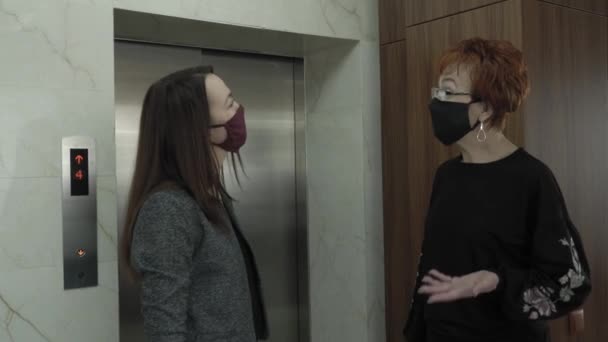 Офис работает во время пандемии COVID-19. Молодая деловая женщина и пожилая женщина в защитных масках разговаривают по офисному коридору и ожидают лифт — стоковое видео