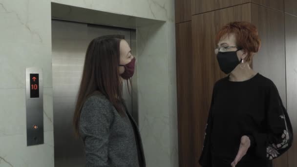 Офис работает во время пандемии COVID-19. Молодая деловая женщина и пожилая женщина в защитных масках разговаривают по офисному коридору и ожидают лифт — стоковое видео