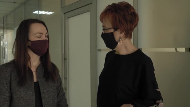 Kontorsarbete under en pandemi COVID-19. Ung affärskvinna och en äldre kvinna i skyddsmasker pratar längs kontoret coridor. — Stockvideo