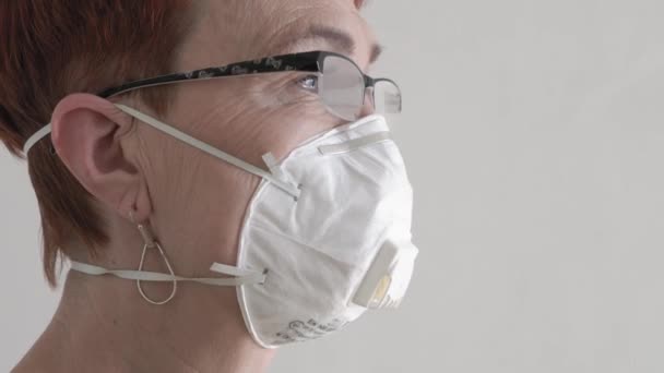 Ηλικιωμένη γυναίκα που φοράει προστατευτική ιατρική μάσκα προσώπου στο σπίτι, καραντίνα από κορωνοϊό, λευκή μάσκα για ενήλικες. — Αρχείο Βίντεο
