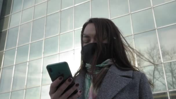 Porträt einer besorgten Frau, die mit Schutzmaske und Smartphone auf der Straße steht. Ein geschäftiges junges Mädchen liest Nachrichten über Coronavirus. Covid-19, Pandemie, Gefahr. — Stockvideo