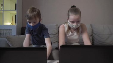 Odaklanmış, koruyucu maskeli tatlı bir çocuk ve kız. İnternette dizüstü bilgisayarda internet kursu çalışması, test sınavına hazırlık, COVID 19 salgını sırasında kanepede otur..