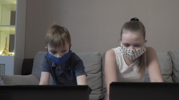 COVID 19 가 유행하는 동안 소파에 앉아서 컴퓨터로 시험 준비를 하는 컴퓨터로 타이핑하는 앱으로 온라인 검색 정보 학습을 하는 귀여운 소년 과 소녀를 집중적으로 보호하는 마스크를 쓴 소년 과 소녀. — 비디오