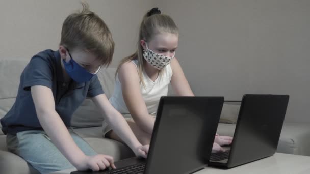 COVID 19 가 유행하는 동안 소파에 앉아서 컴퓨터로 시험 준비를 하는 컴퓨터로 타이핑하는 앱으로 온라인 검색 정보 학습을 하는 귀여운 소년 과 소녀를 집중적으로 보호하는 마스크를 쓴 소년 과 소녀. — 비디오