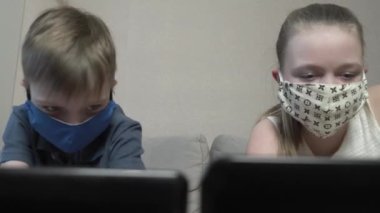 Odaklanmış, koruyucu maskeli tatlı bir çocuk ve kız. İnternette dizüstü bilgisayarda internet kursu çalışması, test sınavına hazırlık, COVID 19 salgını sırasında kanepede otur..