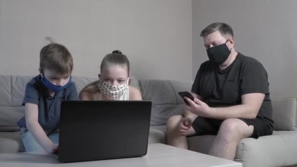 Фокусований милий хлопчик і дівчинка в захисній масці з використанням ноутбука в Інтернеті в додатку, що вводить комп'ютерний іспит, сидить на домашньому дивані під час пандемії COVID-19. Тато зі смартфоном доглядає за дітьми — стокове відео