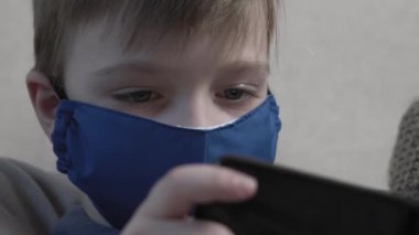 Akıllı telefon kullanarak koruyucu maske takan sevimli çocuk COVID 19 salgını sırasında kanepede oturur..