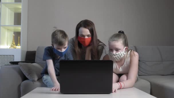 Enfocado lindo chico y chica y su mamá en máscara protectora utilizando el estudio portátil en línea en la aplicación de mecanografía en el examen de ordenador sentarse en el sofá de casa durante la pandemia de COVID 19. — Vídeo de stock