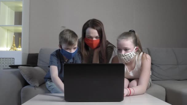 Focalizzato ragazzo carino e ragazza e la loro mamma in maschera protettiva utilizzando lo studio portatile online in app digitando sull'esame del computer sedersi sul divano di casa durante la pandemia COVID 19. — Video Stock