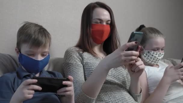 Skupione cute chłopiec i dziewczyna i ich mama w masce ochronnej za pomocą smartfona siedzieć w domu sofa podczas pandemii COVID 19. — Wideo stockowe