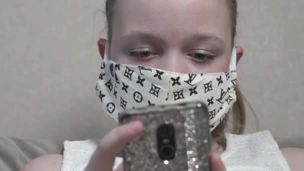 Fokuserad söt flicka i skyddsmask med smartphone sitta hemma soffa under COVID 19 pandemi. — Stockvideo
