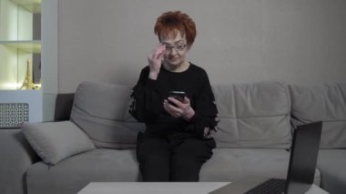 Evde tek başına akıllı telefon kullanan kısa kızıl saçlı yaşlı bir kadın. Dünya çapında küresel COVID-19 salgını.
