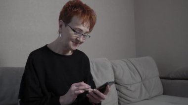 Evde tek başına akıllı telefon kullanan kısa kızıl saçlı yaşlı bir kadın. Dünya çapında küresel COVID-19 salgını.