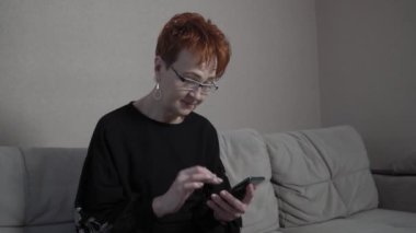 Evde tek başına akıllı telefon kullanan kısa kızıl saçlı yaşlı bir kadın. Dünya çapında küresel COVID-19 salgını.