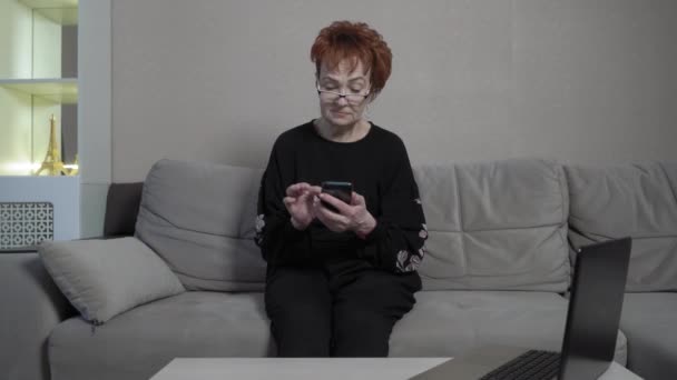 Senior kvinna med kort rött hår hemma ensam med hjälp av smartphone. Världsomspännande epidemi COVID-19. — Stockvideo