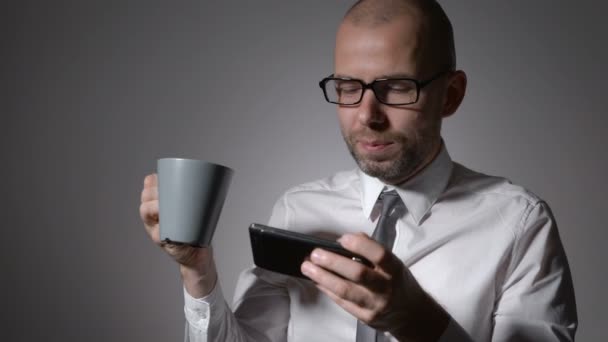 Elegante hombre de negocios con camisa blanca sosteniendo una taza de café o té en las manos. El hombre hizo un descanso en el trabajo, utilizando un teléfono inteligente, para sms o redes sociales — Vídeo de stock