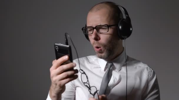 Empresário em uma camisa e fones de ouvido grandes, cantando em um smartphone, usa um aplicativo . — Vídeo de Stock