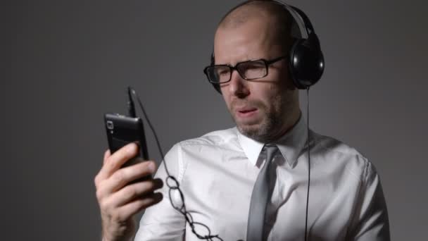 Businessman in a shirt and big headphones, singing in a smartphone, uses an application. — Stock Video