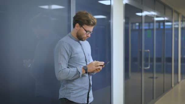 Hombre serio de pie en la pared en una oficina y escribir sms en el teléfono móvil . — Vídeos de Stock