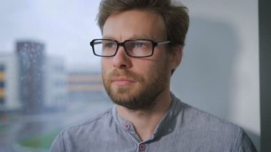 Gözlüklü bir pencere pervazına üzerinde oturan yüzünde dalgın bir ifade ile genç adam.