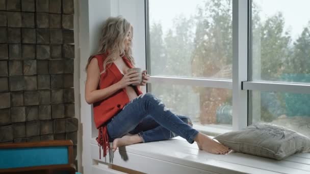 Young pregnant woman in red shirt sitting on the windowsill with the cup of tea. Beautiful future mother drinking tea and thinking about her baby. Future mother smiling and happy. — Stock Video