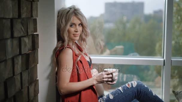 Belle femme blonde enceinte en chemise rouge et jean bleu est assis sur le rebord de la fenêtre près de la fenêtre et le mur avec la tasse de jus. La future mère regarde la caméra , — Video