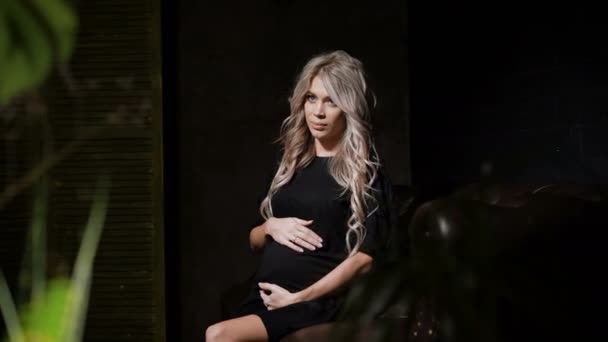 Hermosa mujer embarazada en vestido negro sentada mirando a su izquierda y tocando su vientre amorosamente en la habitación oscura en el gran sofá de cuero marrón con las flores en primer plano. Futura madre sentada en — Vídeos de Stock