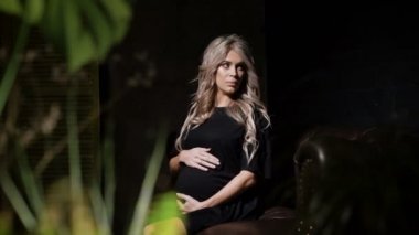 Siyah elbise strocking güzel hamile sarışın karnı sevgiyle çiçek ön plan ile büyük kahverengi deri kanepede karanlık odada oturuyor. Gelecekte anne evde rahatlatıcı.