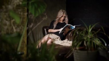 Siyah elbiseli güzel hamile kadın büyük kahverengi deri kanepe ve okuma dergi üzerinde rahatlatıcı. Bekleyen anne evde dönüm sayfaları ön planda çiçekli dergisinin rahatlatıcı.