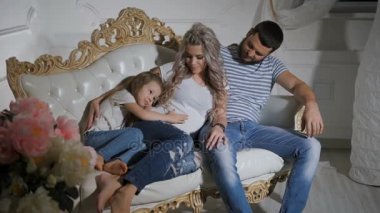 Baba, hamile anne ve küçük kızı birlikte kanepede oturan ve sarılma. Kucaklayan ve okşayarak kızın göbek anneler. Mutlu aile yeni bebek birlikte sarılma bekliyor
