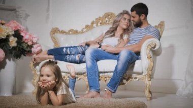 Mutlu bir aile ile gülümseyen kız ön planda, yeni bebek bekliyor, Close-Up halı üzerinde yalan, bacaklarını sarkan ve ellerini sallayarak.