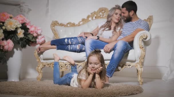 Primer plano de la familia feliz esperando un nuevo bebé juntos en la habitación. Hay una pequeña hija en primer plano, acostada en la alfombra, colgando las piernas y sonriendo . — Vídeos de Stock