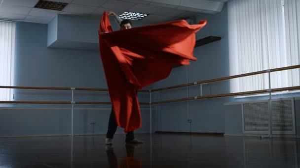 The young man in the rehearsal hall. Rehearses dance for statement. He by means of red fabric does rotations. Fabric gives to dance grace. — Stock Video