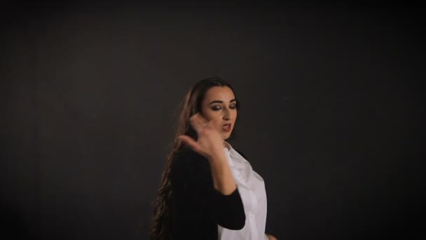 Стильна довгошерста молода жінка в білій сорочці і чорній куртці виконує сучасні джазові танці з руками і тілом з чорною стіною на задньому плані . — стокове відео