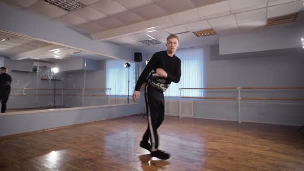 在黑色的裤子和套衫在教室里表演霹雳舞的青年舞蹈家。他正在他的胳膊和腿展示现代舞蹈。少年看起来像专业舞蹈演员. — 图库视频影像