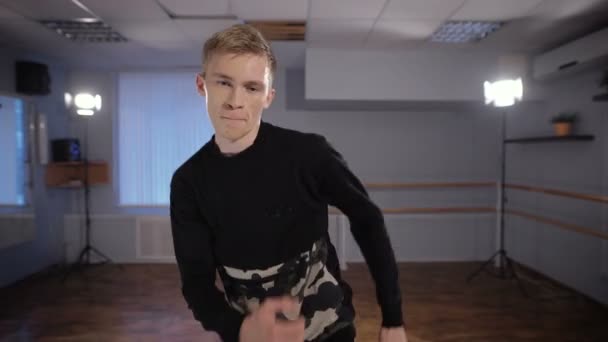 Självsäker hip hop dansare aktivt dans och tittar in i kameran i en dansstudio. Ung man utför danssteg med händerna. — Stockvideo