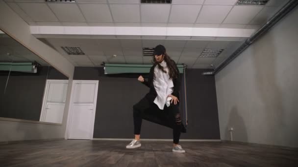 Kobieta w puste studio bierze lekcje tańca. Ubrana jest w bezpłatne koszulę w Pelerynie, na głowie jej czapkę. W hali ona jest skoncentrowany spełnia hip-hop ruchu.. — Wideo stockowe