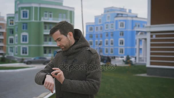 Domy Courtyard. Mężczyzna w szarym płaszczu z zegarem z strony i z pierścieniem na palcu jest na ścieżce. On zmienia ustawienia w zegarku i raduję się, on robi wielkie. — Wideo stockowe