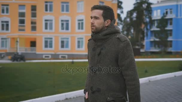 Homem sem barba, alto a caminhar pelo caminho. Ele usava um casaco cinzento. Ele é focado e sério. Casa de cores laranja, azul e marrom. No quintal gramado verde Paisagado — Vídeo de Stock