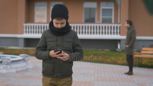 Boy v teplé tmavě šedou bundu a černou čepici a šálu používá telefon stojící na ulici se svým otcem za ním. Manžel a syn čekají na své manželky a matky — Stock video