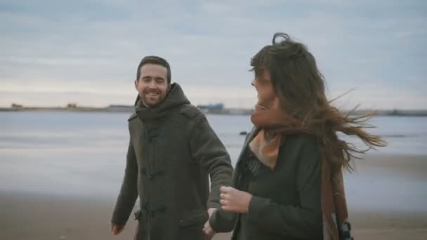 幸せなカップルは、一緒に手を取り合って砂の海岸に沿って実行しています。黒いコートと茶色のスカーフの若い女性は笑って、彼女の夫と共に実行されます。. — ストック動画
