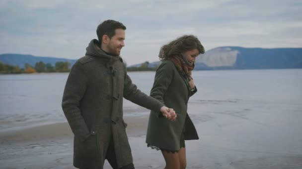 Coppia romantica correre e saltare insieme lungo la riva del mare. Le persone felici si stanno tenendo per mano. Giovane donna in cappotto nero e sciarpa marrone sta ridendo e correndo insieme — Video Stock