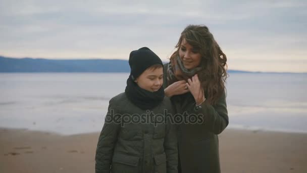 Närbild av mor och son i varma kläder står på stranden tillsammans med havet bakom. Mor säger något till sonen inställning sin halsduk, då hon omfamnar honom. — Stockvideo