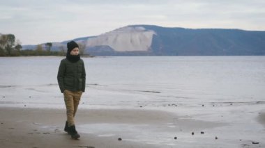 Derin gri ceket, hardal pantolon, siyah şapka ve atkı yalnız sersem arkasında harika büyük Dağları ile sahil kıyısında yürüyüş şık genç.
