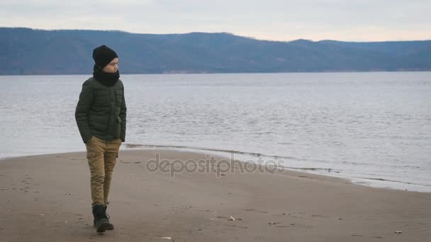 Niño con chaqueta, sombrero negro y una bufanda en el cuello camina por la playa. En el contexto de una visión. Hermoso mar, cautivadoras montañas, olas bajas . — Vídeos de Stock