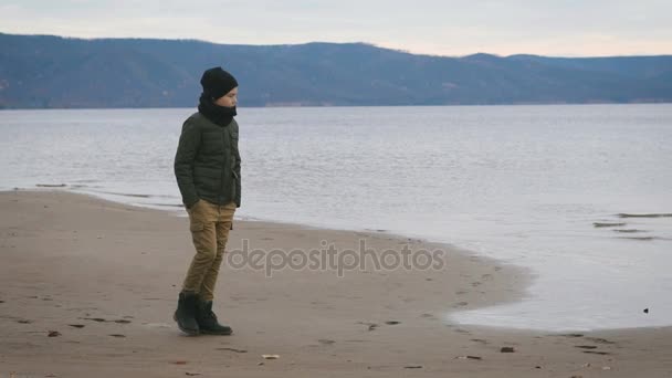 暖和的衣服的男孩正沿着海岸的海滨出神地看着水与山中的背景。时尚少年身着深灰色夹克，芥末裤子, — 图库视频影像