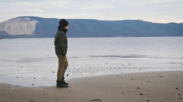 Menino vestido calorosamente passeia ao longo da praia. É casaco quente, chapéu, cachecol, calças e botas cor Lightstone. No contexto de uma visão. Mar bonito, montanhas cativantes, onda baixa . — Vídeo de Stock